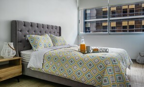 Ein ordentlich bezogenes Bett mit gemusterten Bettwaren, ein Nachttisch mit Lampe und ein Fenster mit Jalousien.