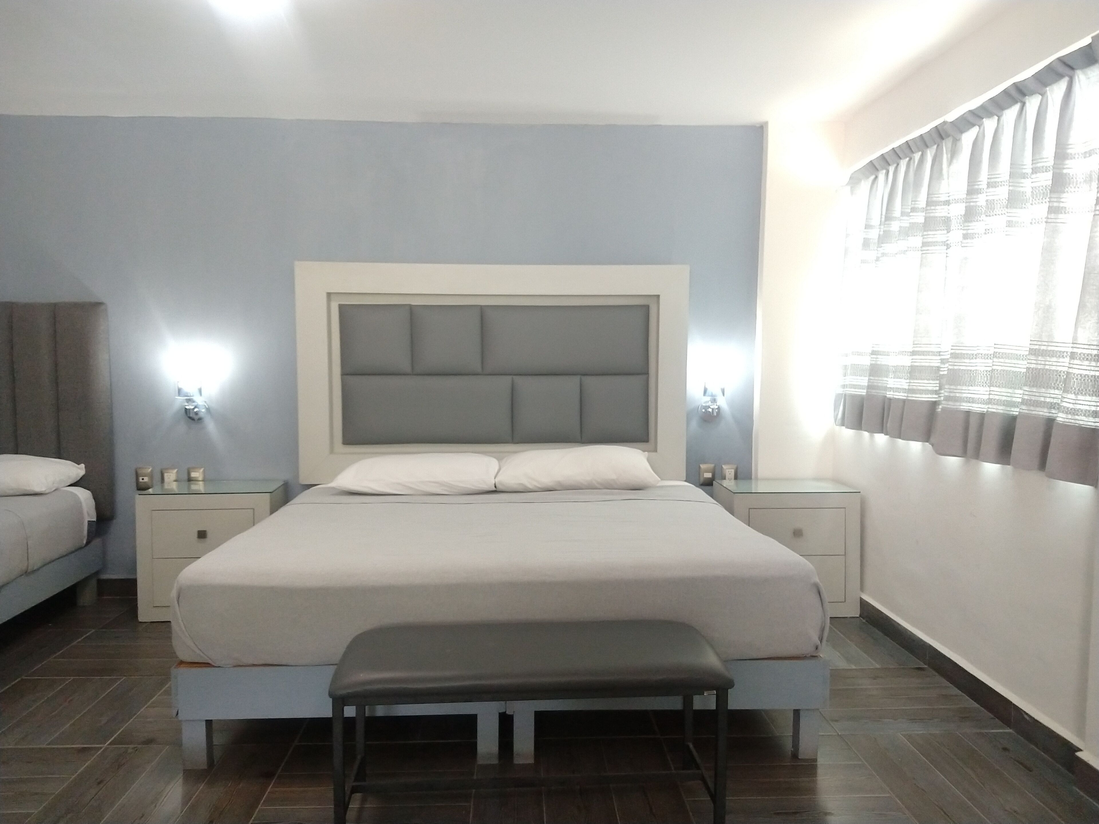 A modern bedroom with a large bed, bedside tables, a bench, and a window with curtains.