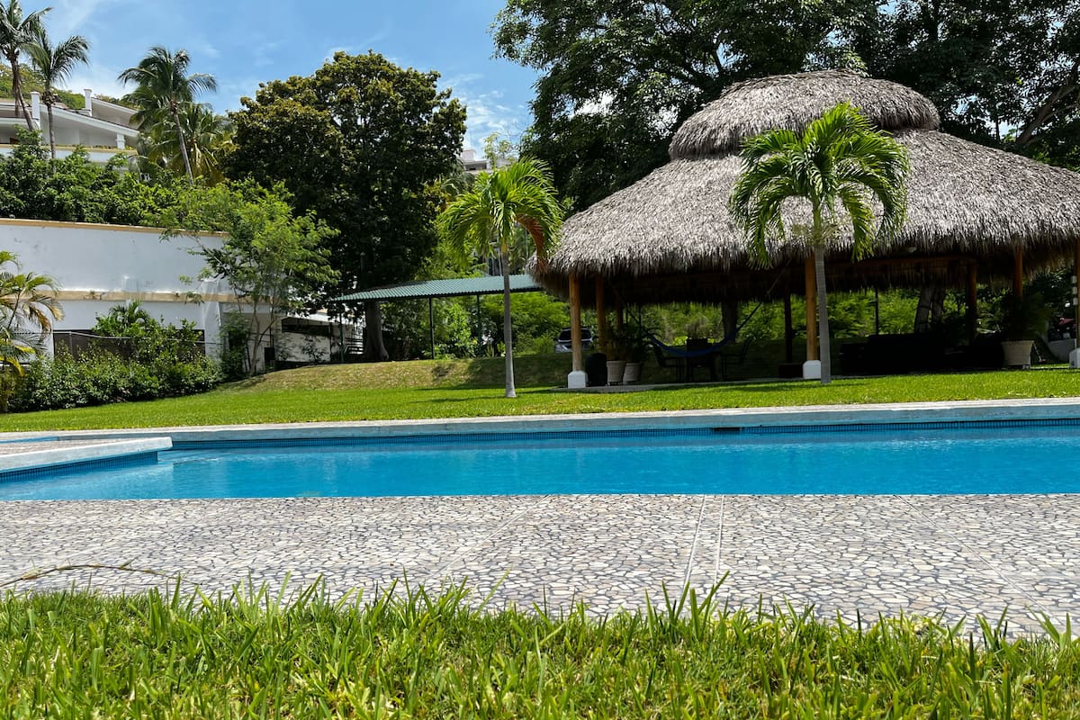Una piscina con una estructura de techo de paja, rodeada de exuberante vegetación y un edificio blanco al fondo.