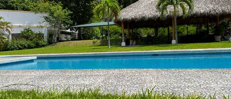 Una piscina con una estructura de techo de paja, rodeada de exuberante vegetación y un edificio blanco al fondo.