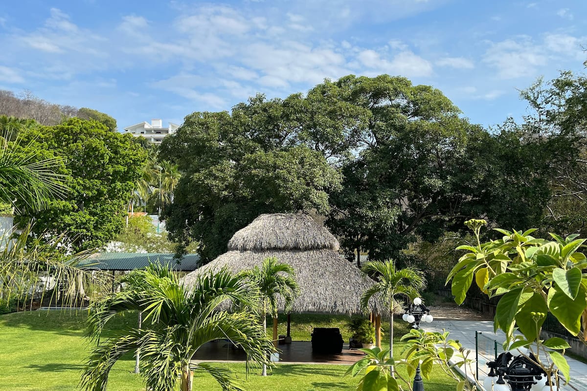 Una piscina rodeada de exuberante vegetación, una estructura con techo de paja y un cielo azul cristalino.