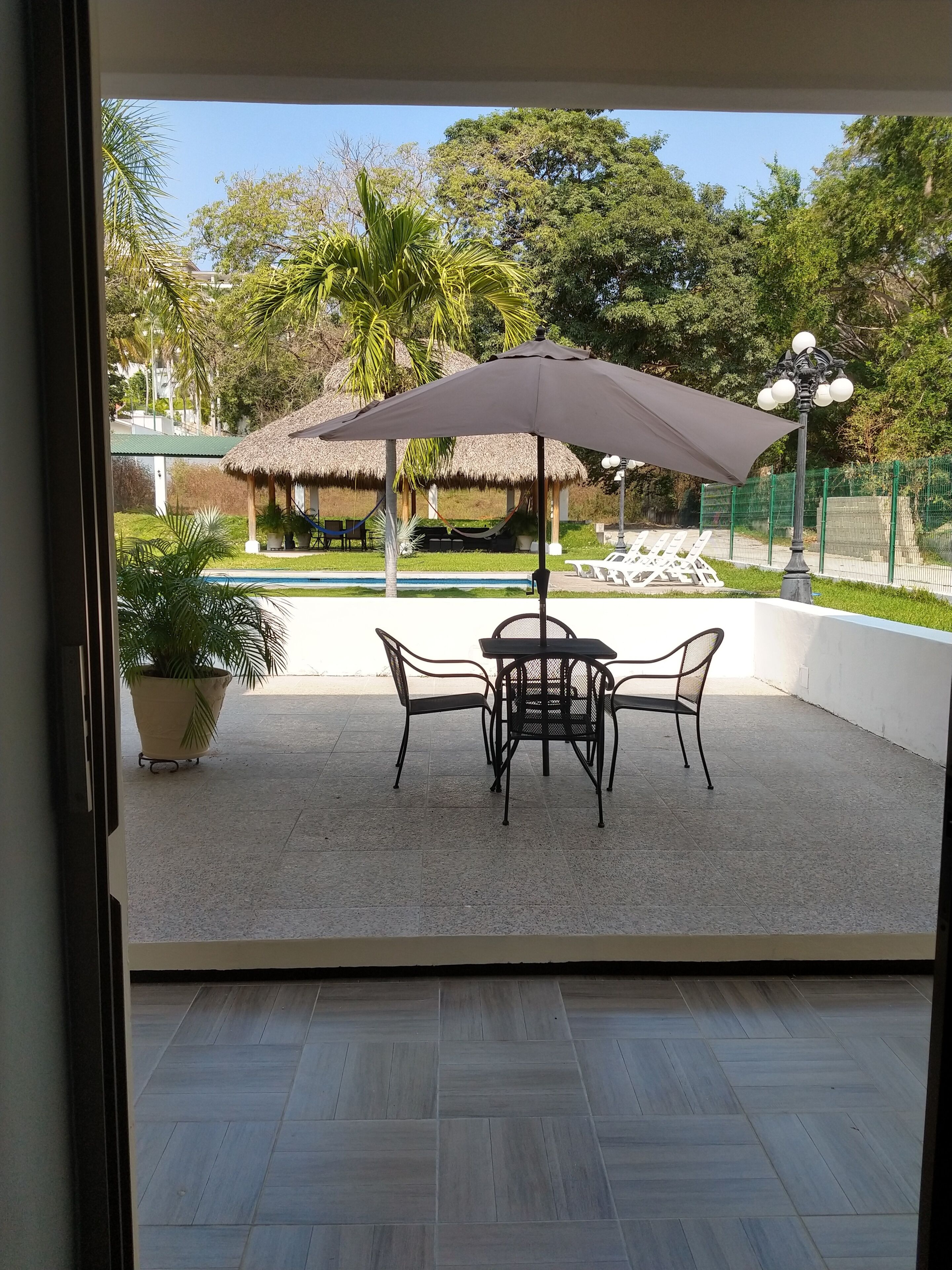 A patio with a table and chairs under a large umbrella, a potted plant, and a view of a pool and thatched-roof structures.