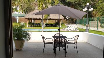 Un patio con mesa y sillas bajo un gran paraguas, una planta en maceta y vistas a una piscina y construcciones con techo de paja.