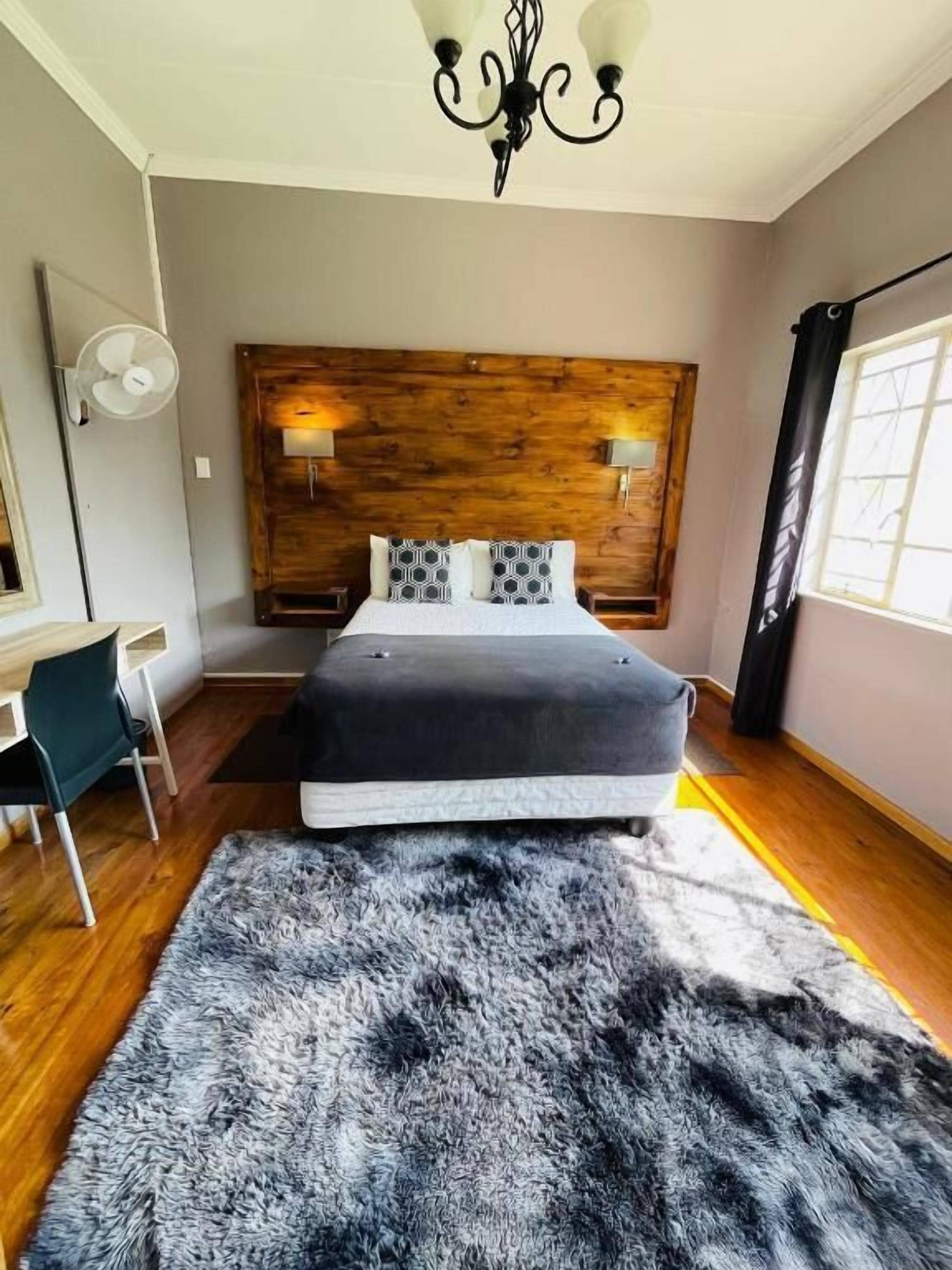 Een slaapkamer met een houten hoofdbord, een bed met een grijze matras, een zwart-wit tapijt, een bureau met een stoel en een raam met gordijnen.