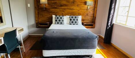 A bedroom with a wooden headboard, two bedside lamps, a bed with a grey blanket, a desk with a chair, and a window with curtains.