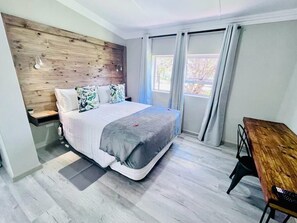 A modern bedroom with a wooden headboard, a large bed with a grey and white duvet, a bedside table, and a wooden bench. - Madeira Bed (Komani)