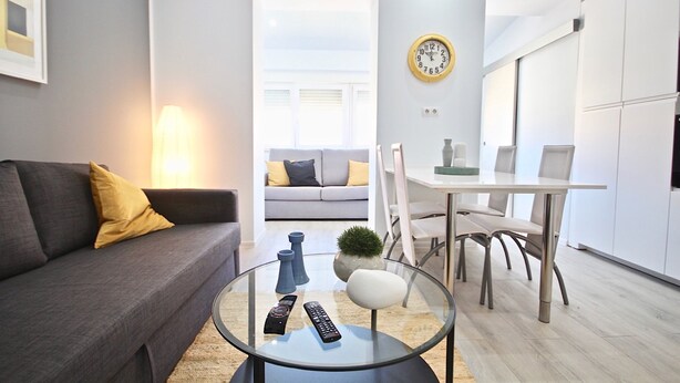 A modern living room with a grey sofa, a glass coffee table, and a dining area with a white table and chairs.