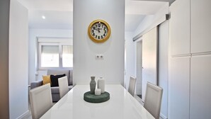 A modern dining area with a white table, a clock on the wall, and a sofa in the background.