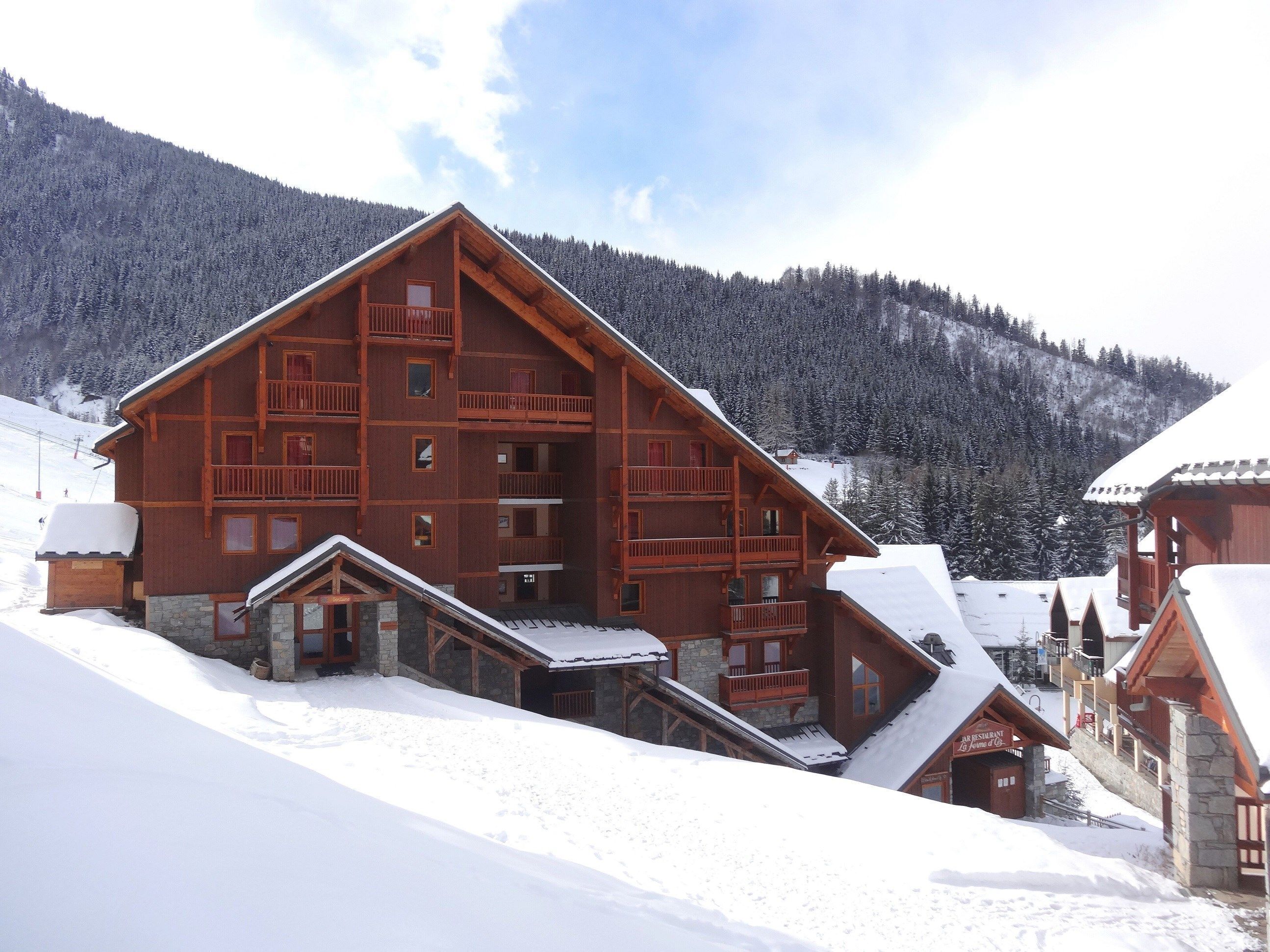 A large wooden chalet with multiple balconies and a stone foundation, surrounded by snow-covered mountains.