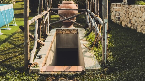 Une structure rustique en plein air, dotée d’une ossature en bois et d’un grand pot en terre cuite placé au centre, le tout se dressant sur un fond de pelouse et d’une colline rocheuse.