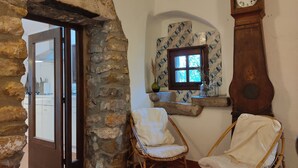 Un arco de piedra que da acceso a una habitación con sillas de mimbre, un reloj y una ventana con baldosas estampadas.
