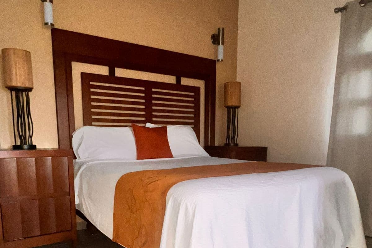 A hotel room with a wooden headboard, a bed with white and orange bedding, two bedside lamps, and a wooden nightstand.