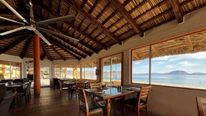 A restaurant with a thatched roof, wooden furniture, and large windows offering a view of the sea. - Los Vientos Hotel (Bahia de los Angeles)