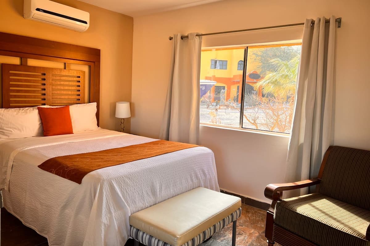 A hotel room with a bed, a bench, a window with curtains, and an air conditioning unit.