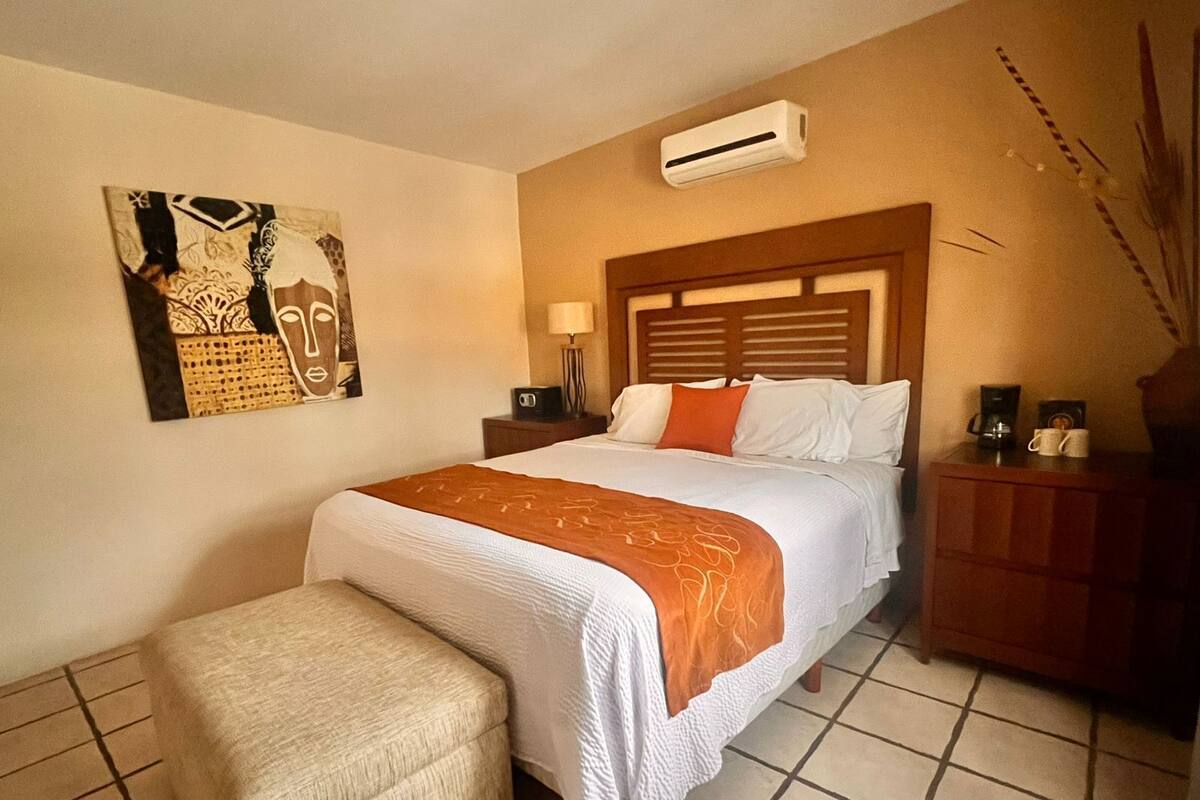 A hotel room with a bed, a nightstand, an air conditioner, a painting on the wall, and a small table with a vase.