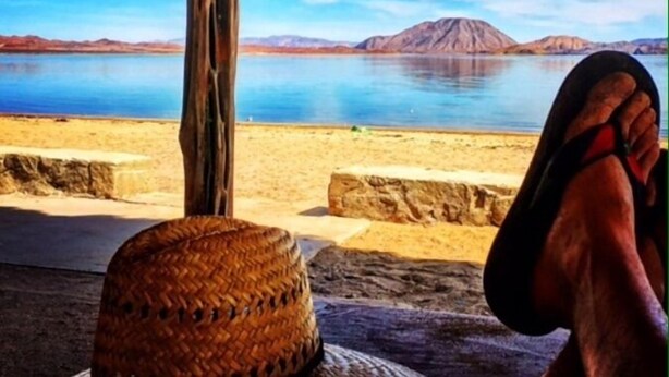 A beach view with a thatched roof structure, a woven basket, and a person relaxing with a mountain in the background. - Los Vientos Hotel (Bahia de los Angeles)