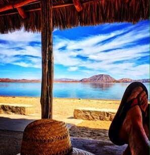 Vista de playa con una estructura de techo de paja, una cesta tejida y una persona relajándose con una montaña al fondo.