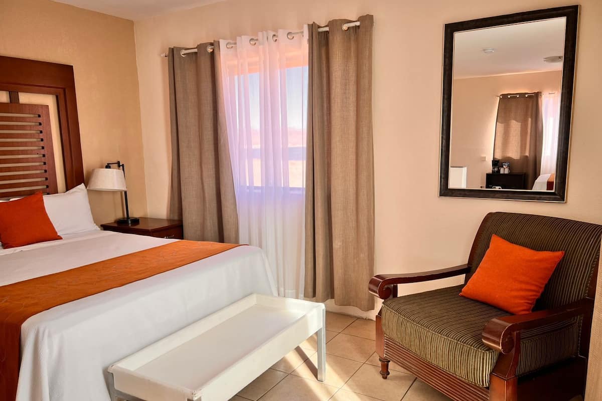 A hotel room with a bed, a chair, a bench, a nightstand, a lamp, a window with curtains, and a mirror on the wall.