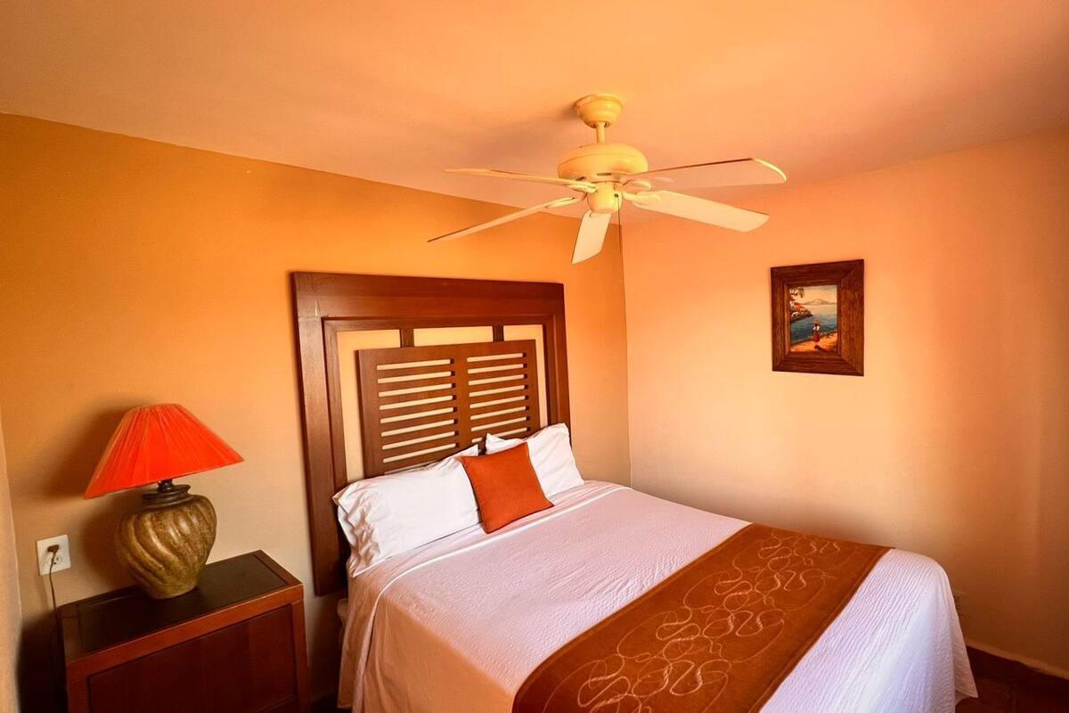 A hotel room with a bed, a nightstand, a lamp, a ceiling fan, and a painting on the wall.