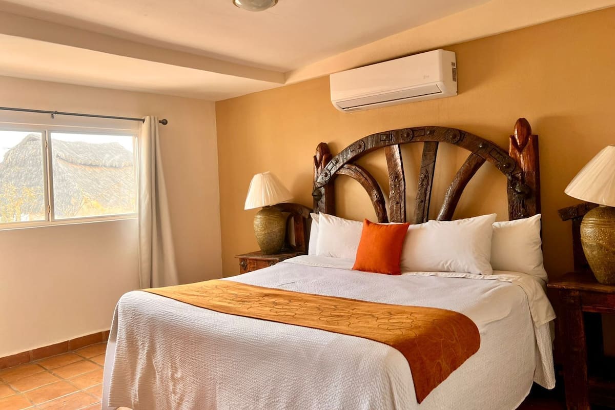 A hotel room with a bed, two bedside lamps, an air conditioning unit, a window with curtains, and a view of the outside.