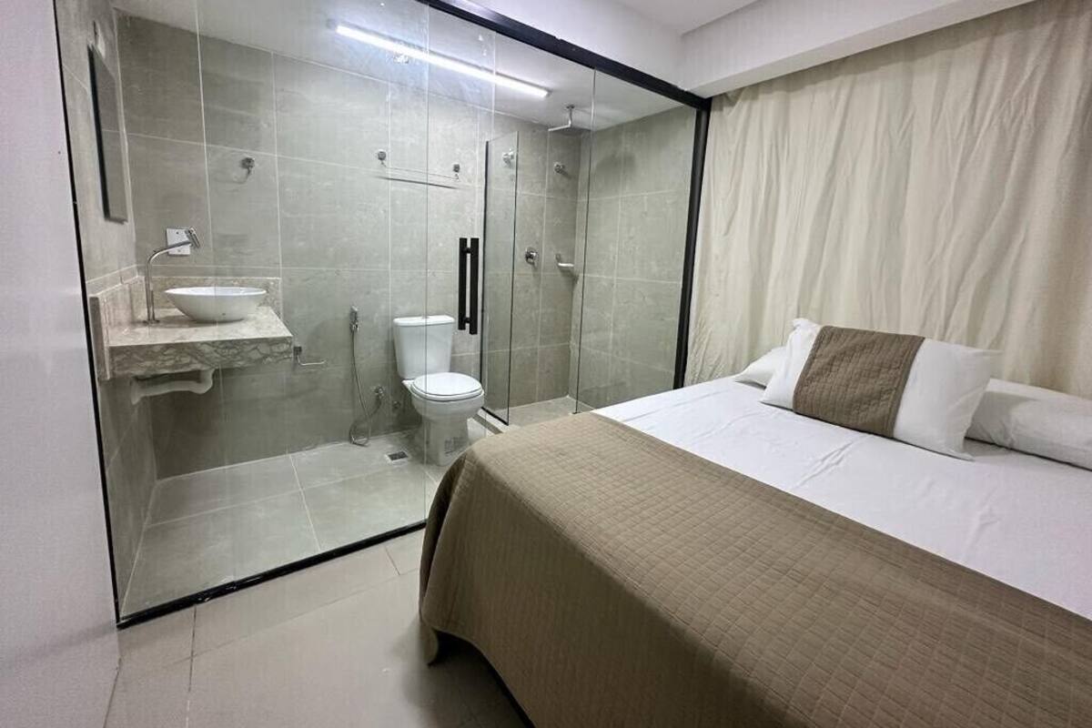Une chambre d’hôtel moderne dotée d’une douche en verre, d’un lit recouvert d’une courtepointe beige et d’une salle de bain blanche avec un lavabo et des toilettes.