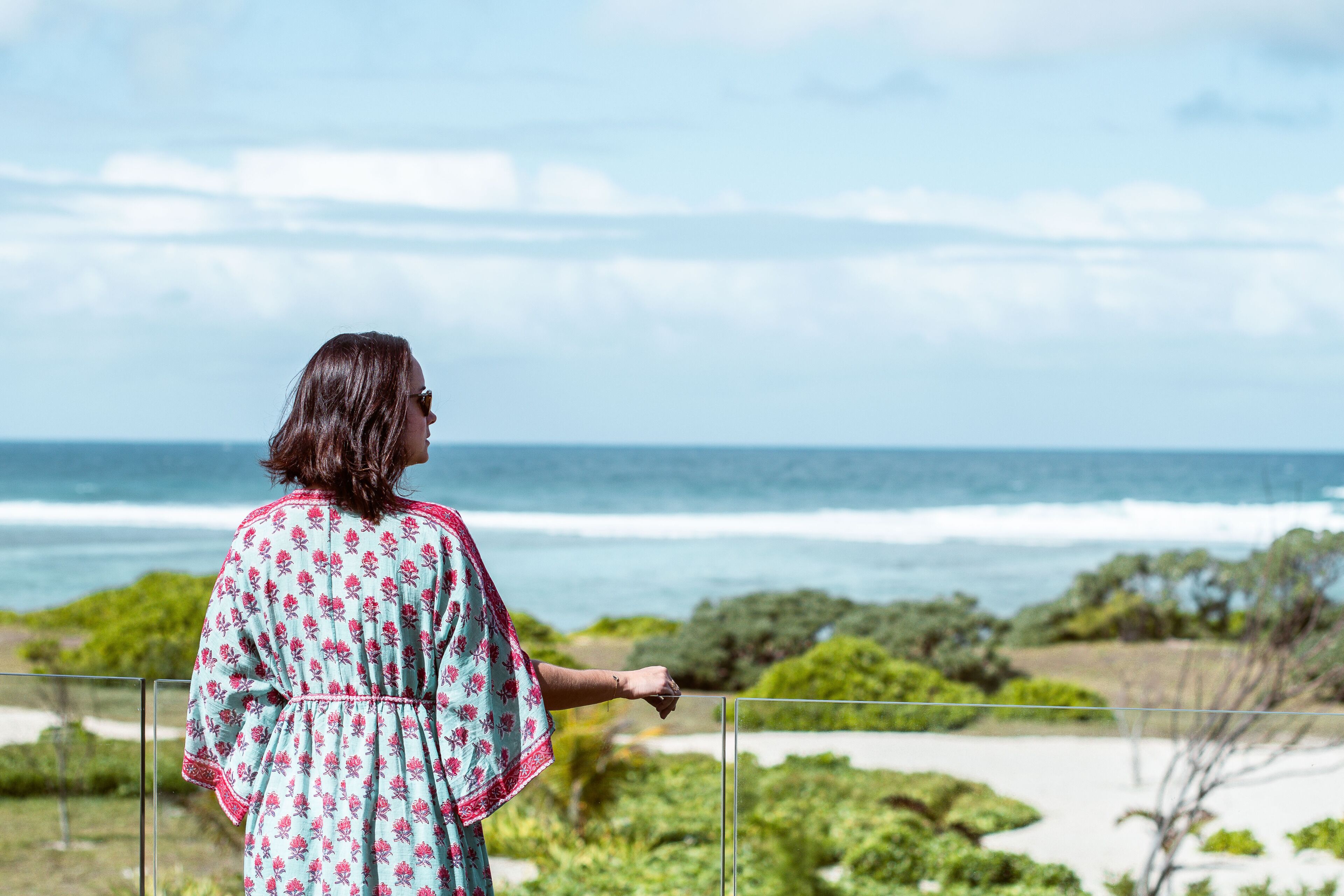 Une personne se tient sur un balcon donnant sur une plage, avec l’océan, ses vagues et la végétation environnante.