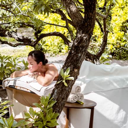 A person relaxing in a hammock under a tree, with a towel and rolled towels on a small table nearby.