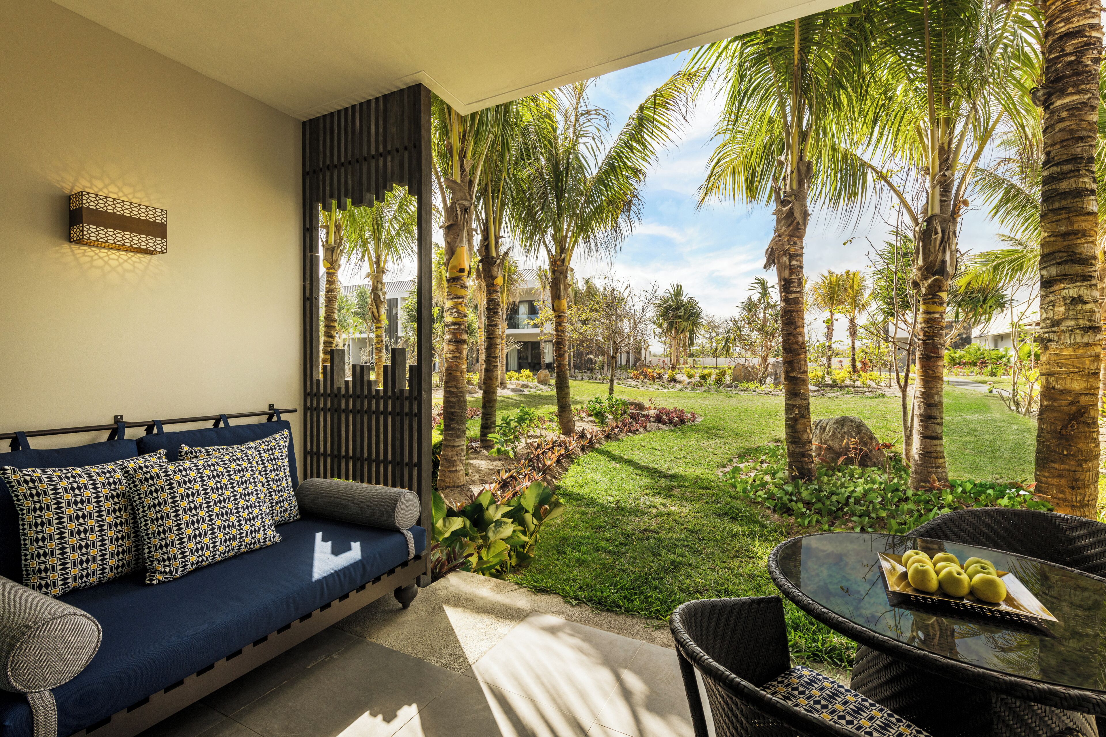 A modern living room with a sofa, a dining table with chairs, and a view of a tropical garden with palm trees.