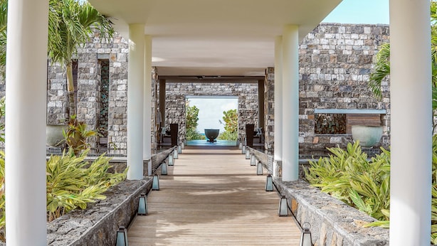 A wooden pathway leading to a stone wall with a large bowl in the center, flanked by columns and plants. - Constance Le Chaland Iko Mauritius (Blue Bay)