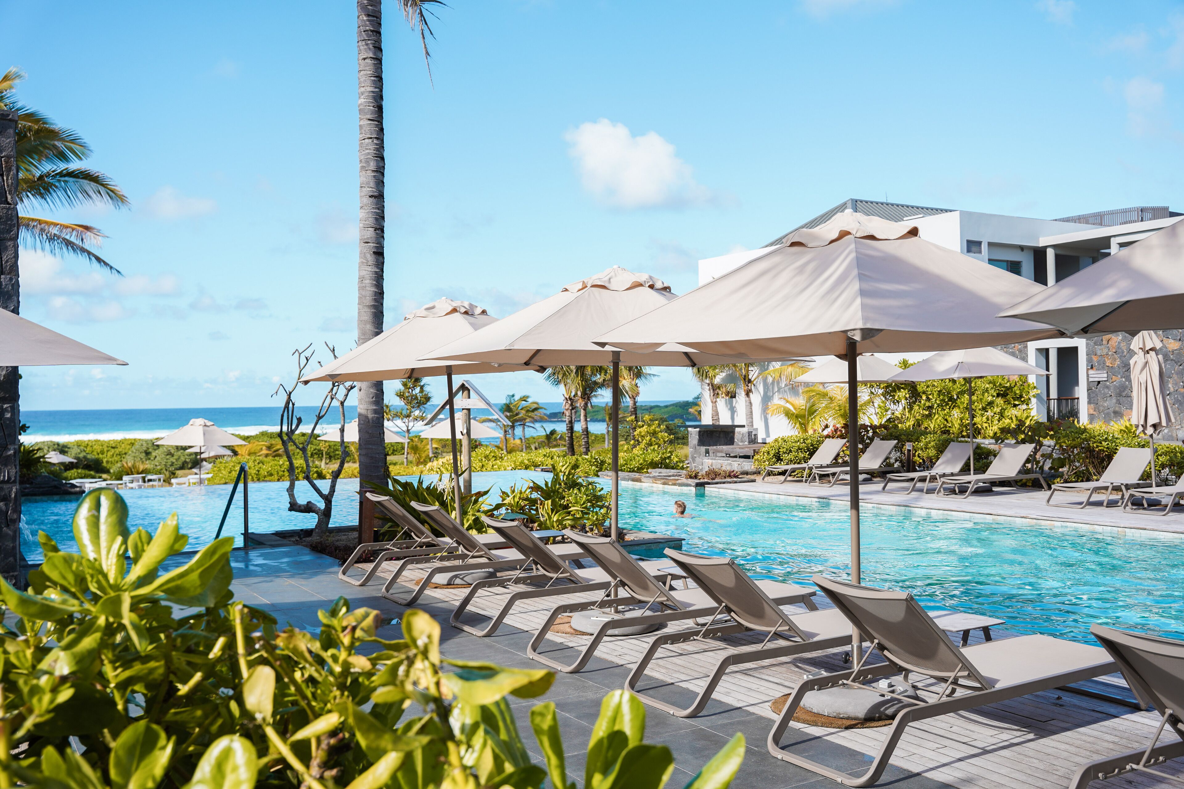 A pool area with lounge chairs and umbrellas, a clear view of the ocean, and a modern building in the background.