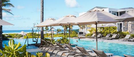 A pool area with lounge chairs and umbrellas, a clear view of the ocean, and a modern building in the background.