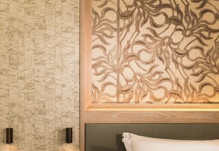 A hotel room with a bed, a nightstand with two vases, a textured wall, and a decorative panel above the bed.