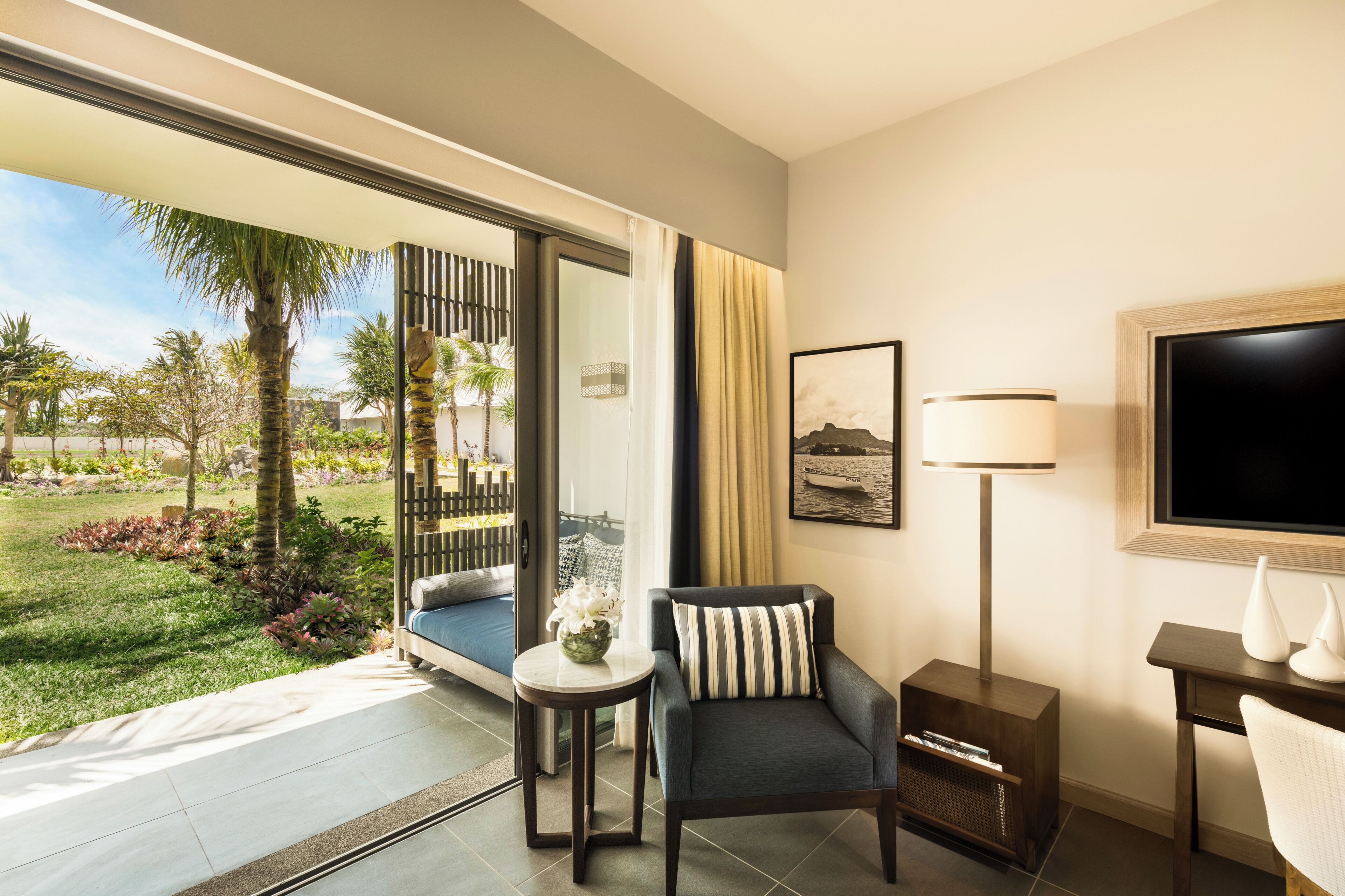 Ein Hotelzimmer mit einer großen Schiebeglastür, die zu einem Garten führt, einem Sofa, einem kleinen Tisch mit einer Vase, einer Lampe und einem Fernseher an der Wand.