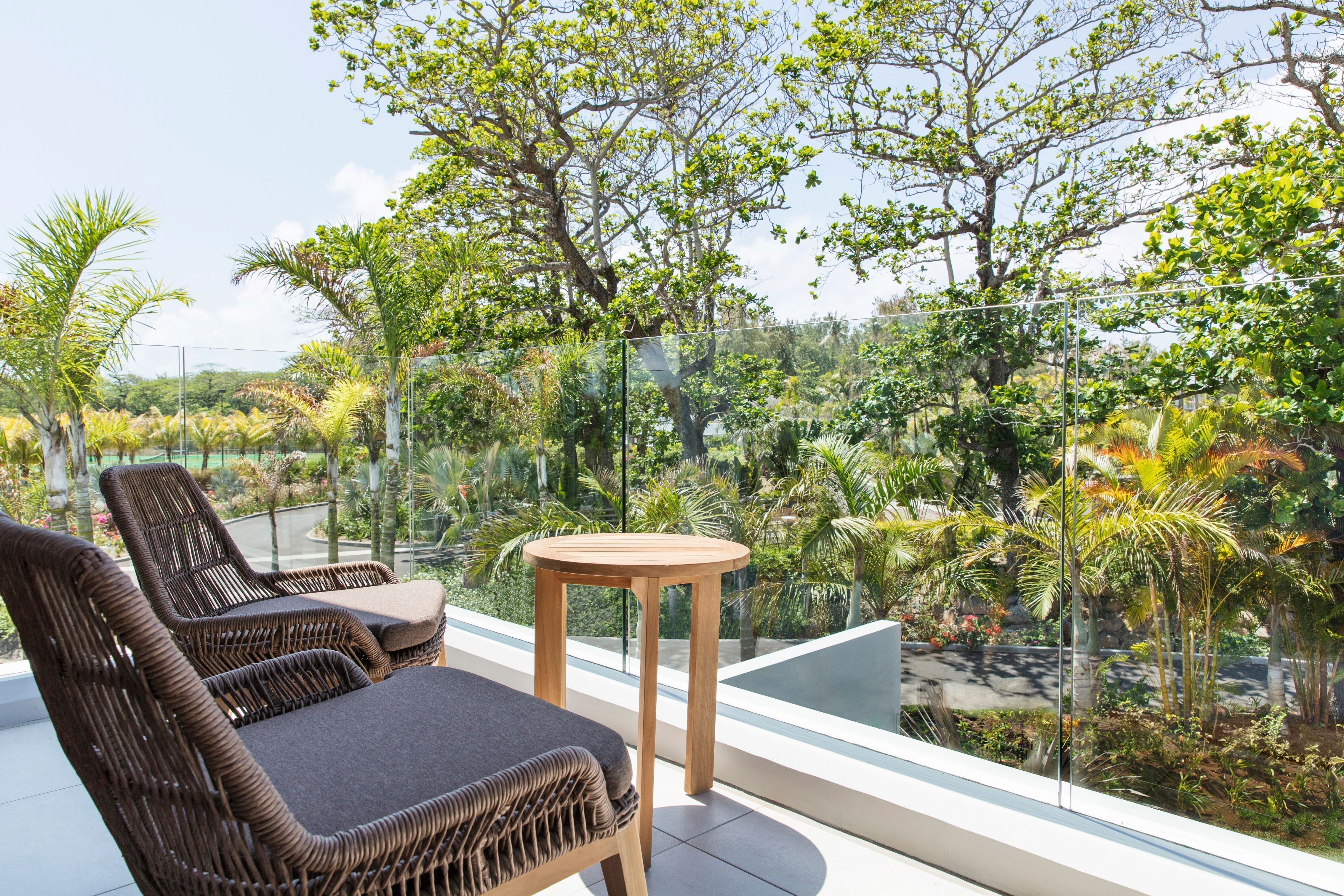 Un balcón con sillones de mimbre y una pequeña mesa de madera, con vista a un jardín exuberante y un cielo despejado.