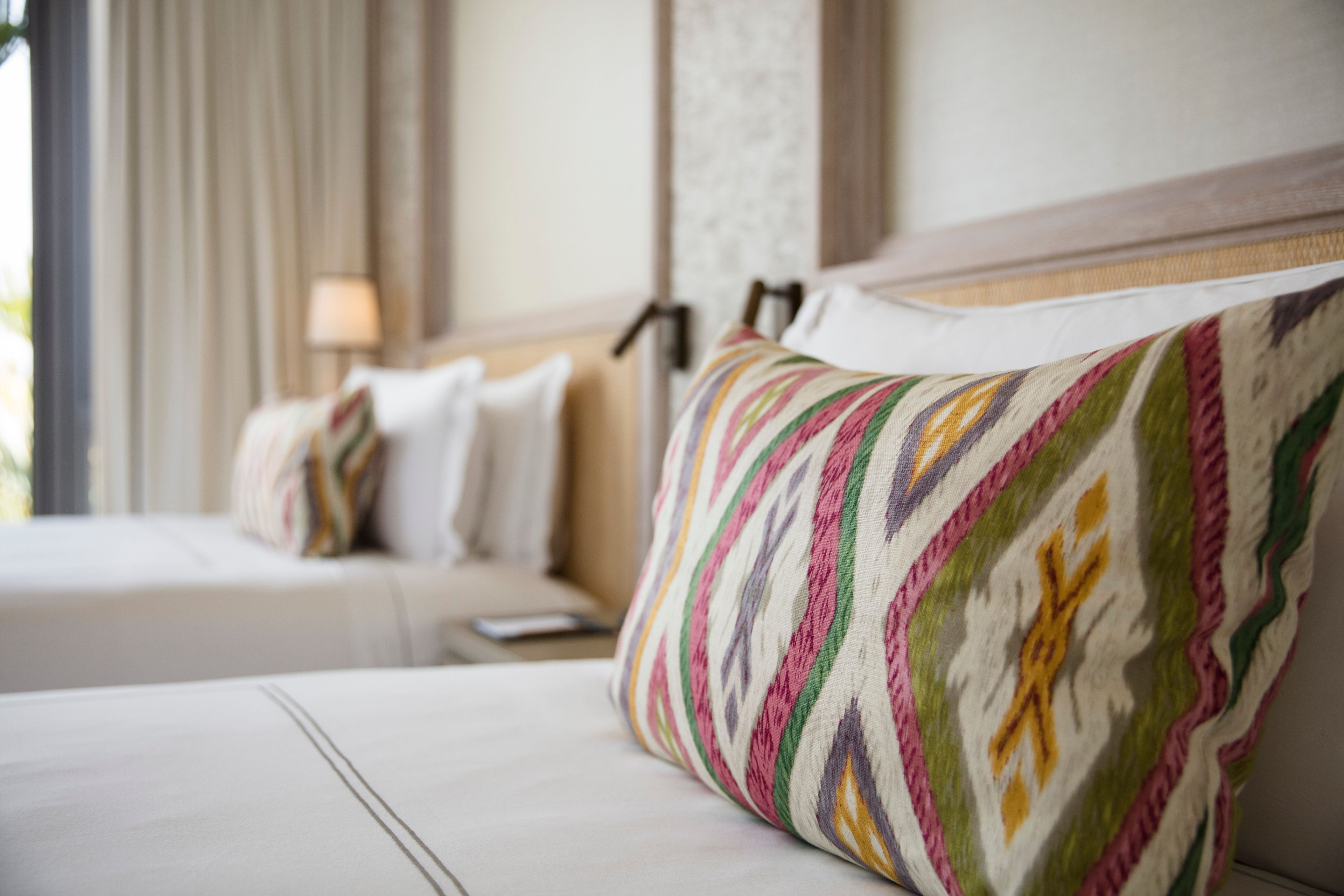 A hotel room with a neatly made bed, a colorful pillow, and a lamp on the nightstand.