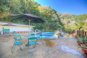 Zona de piscina al aire libre con sillas y mesas, rodeada de abundante vegetación.