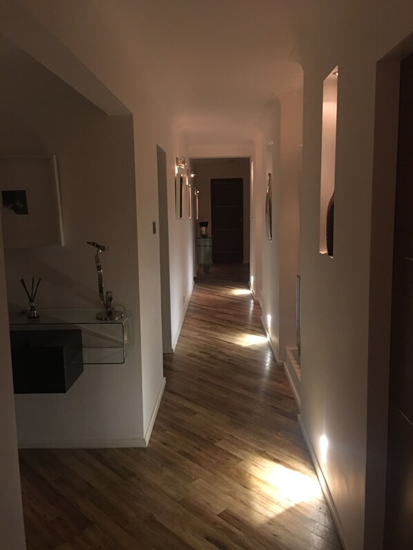 A narrow hallway with wooden flooring, white walls, and recessed lighting.