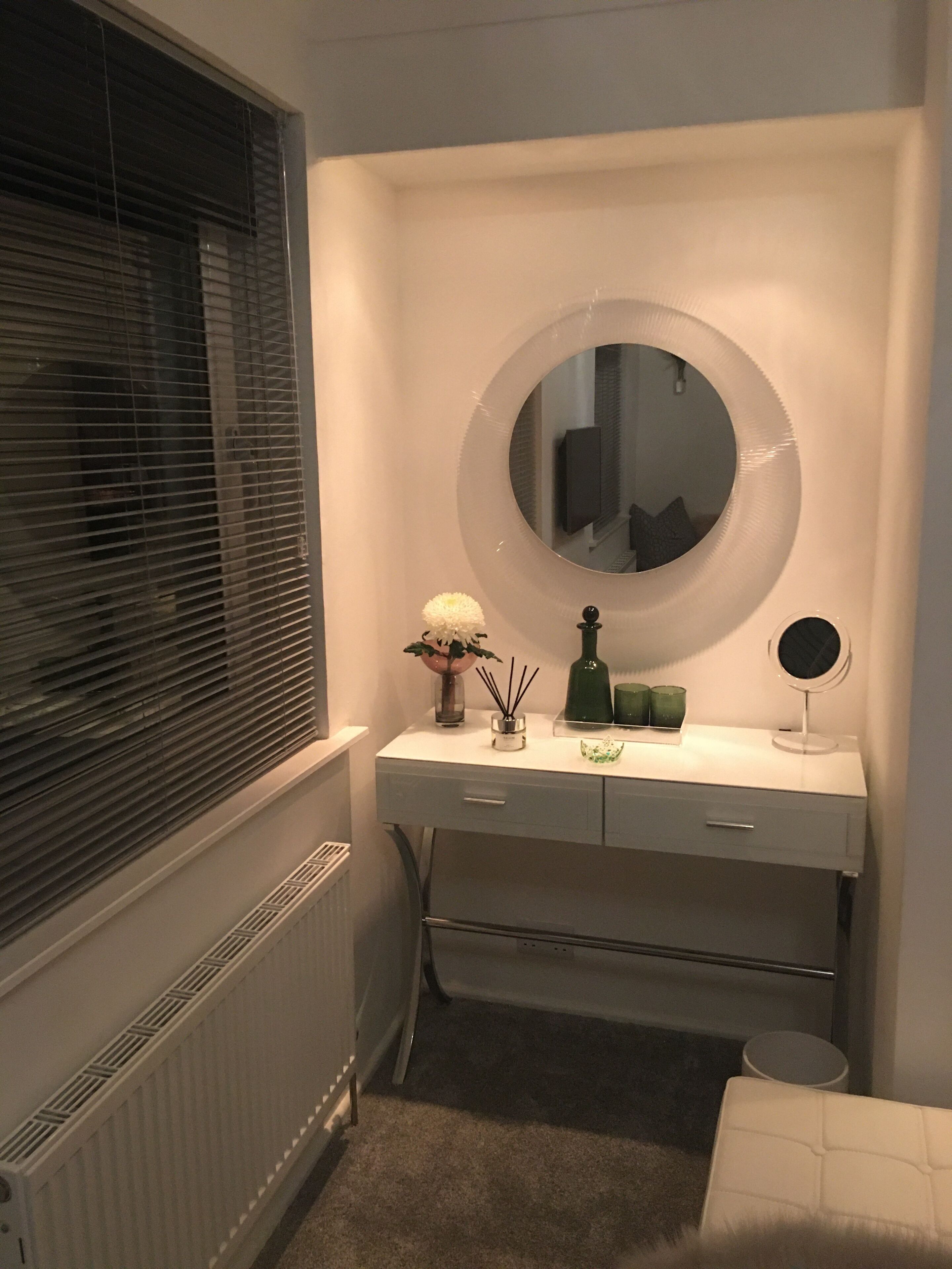 Un bagno con un mobile lavabo bianco, uno specchio rotondo, un piccolo specchio tondo sul mobile lavabo, una bottiglia verde, un vaso bianco con un fiore e un termosifone.
