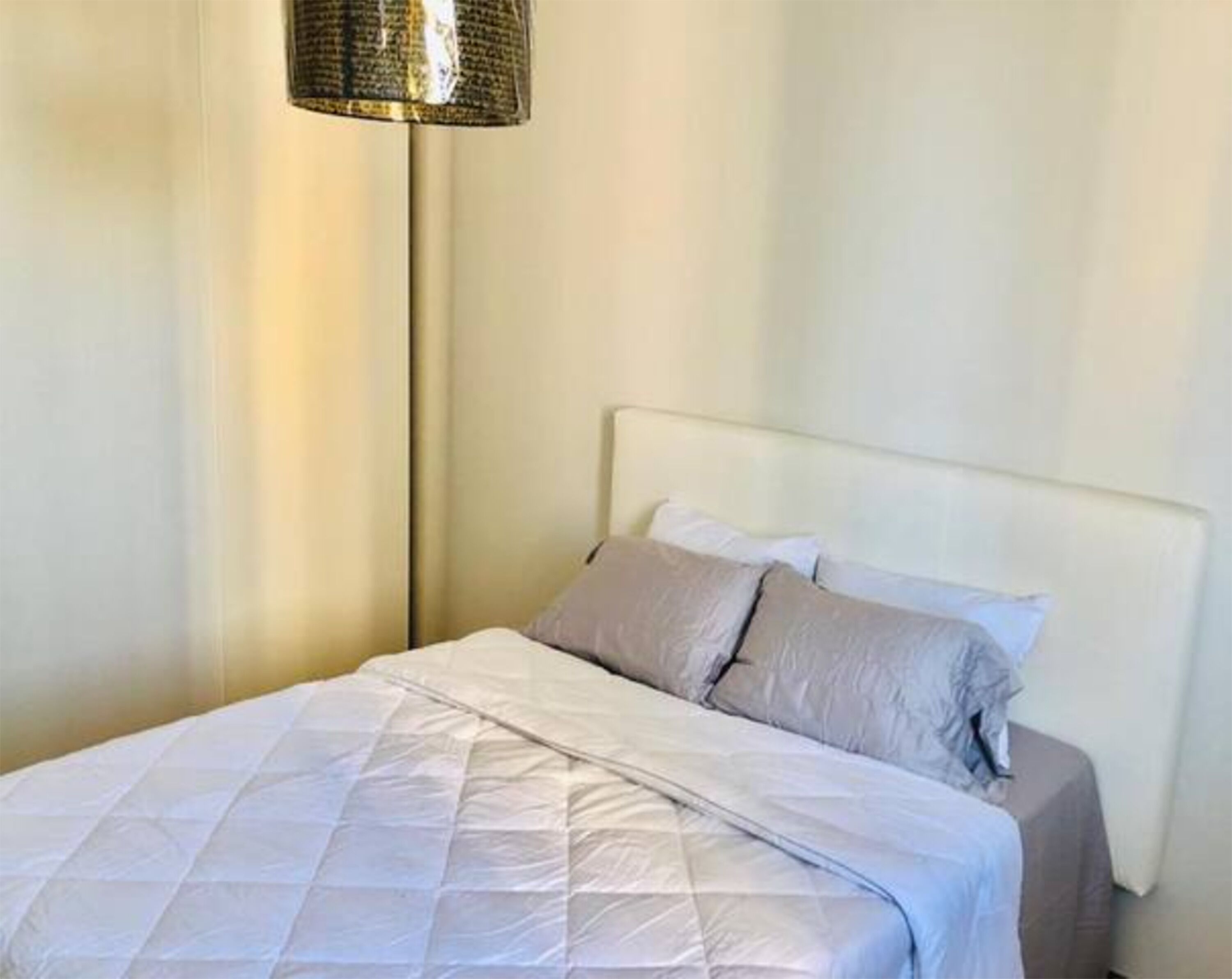 Cama bem arrumada com colcha branca e dois travesseiros, luminária de cabeceira e parede lisa.