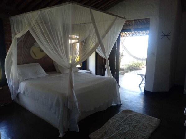A room with a bed under a canopy, a small table, and a view of the outside. - Casarao Alto Mucuge (Porto Seguro)