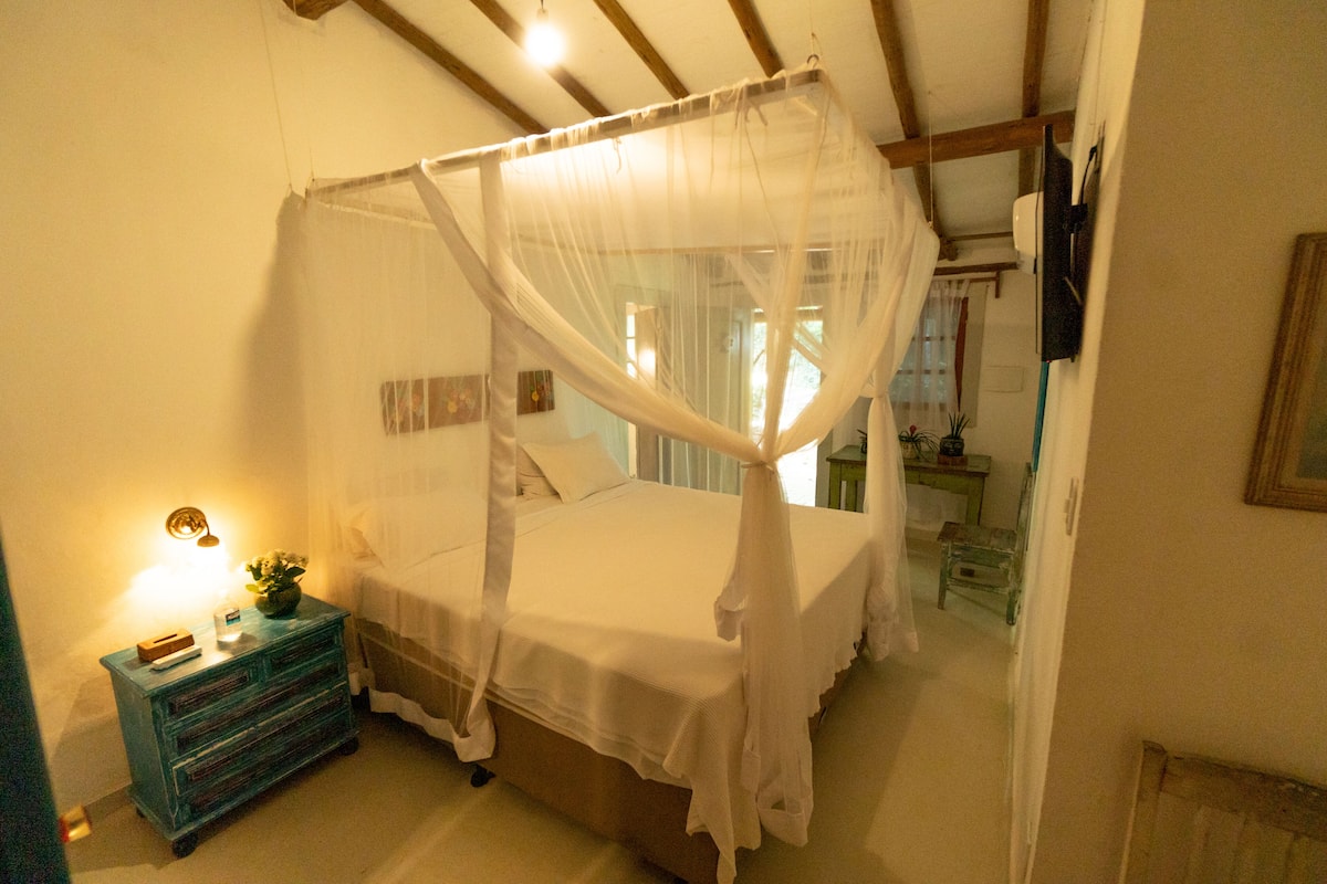 A bedroom with a four-poster bed, a nightstand with a lamp, and a small table with a plant.