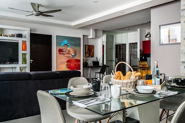 Un salon moderne avec une table à manger en verre pour deux personnes, un canapé noir et un tableau accroché au mur.