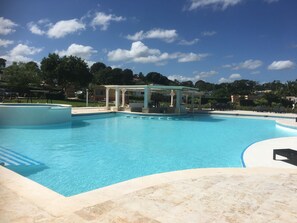 A large outdoor swimming pool with a covered area, surrounded by a landscaped garden and buildings in the background. - Coco Bohio, Villas in Sosua, Casa Linda (Sosúa)