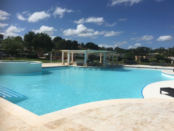 A large outdoor swimming pool with a covered area, surrounded by a landscaped garden and buildings in the background. - Coco Bohio, Villas in Sosua, Casa Linda (Sosúa)