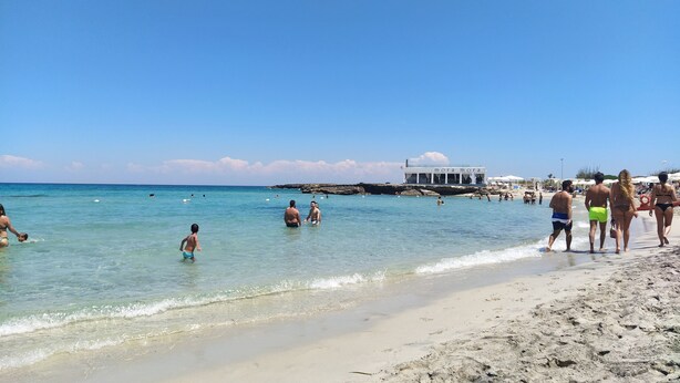 Ein Strand mit klarem Wasser, Menschen beim Schwimmen und Spazierengehen am Ufer, und ein Gebäude mit der Aufschrift 'mora mora' im Hintergrund.
