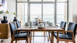 Un espace repas avec une grande table dressée pour six personnes, entourée de chaises, et offrant une vue sur l’océan grâce à de grandes fenêtres.