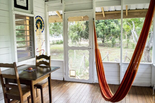 A wooden porch with a hammock, a table with a chair, and a door with a view of trees.