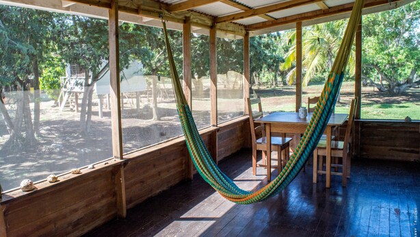 A wooden deck with a hammock, wooden table, and chairs. - Backpackers Paradise (Sarteneja)