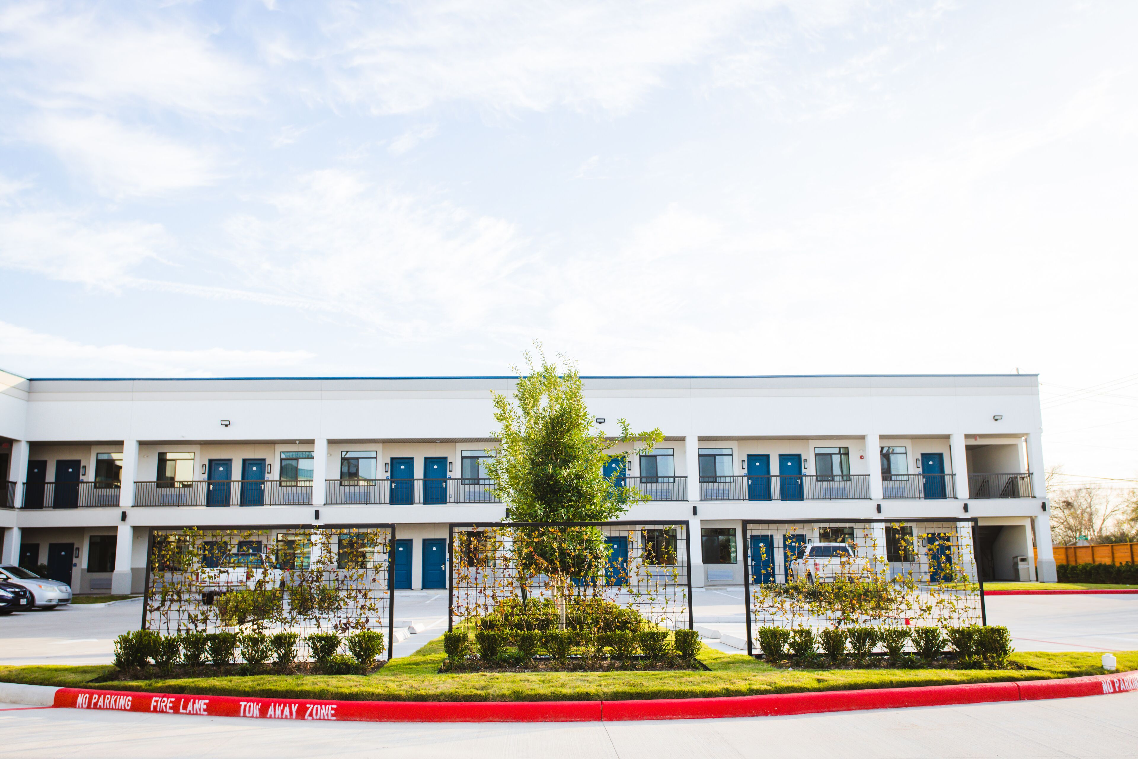 A modern multi-story building with a flat roof, multiple balconies, and a central landscaped area with a tree and shrubs.