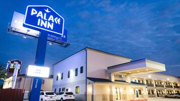 A hotel named "Palace Inn" with a "Now Hiring" sign, a parking lot with several cars, and a modern building facade.